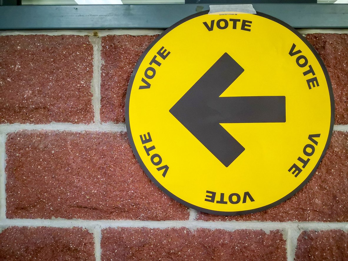 Image of a yellow circle with vote on it and an arrow pointing to the left direction against a brick wall